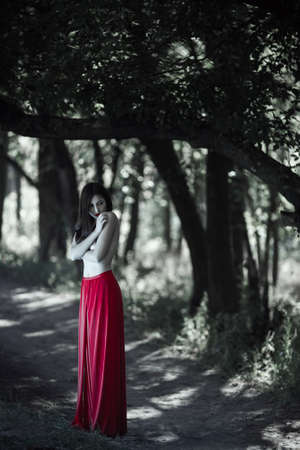 Wonderful sexy fashion model with naked walking in a fantastical forest. Photo of seductive nude woman in full length in luxury long red dress posing against bokeh trees background. Multi-racial Asian Caucasian girl. Fashionable toning. Creative computer の写真素材