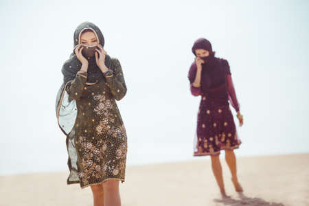 Desert women thirsty dehydrated tired walking outdoors. Dehydration, overheating, thirst and heat stroke concept image with two sisters in desert nature. Beautiful mixed race asian caucasianl arabian girls lost in desert during journey.の写真素材