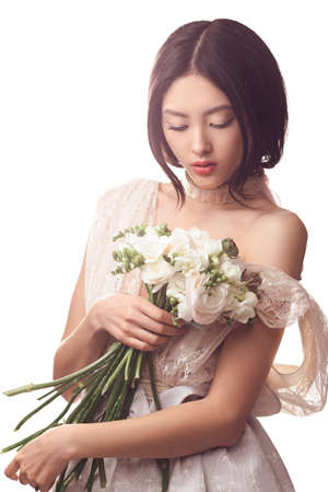 Bride. Woman in the lacy white dress with flowers peonies in hands on a light background. Asian female model posing in studio with bouquet of spring flowers.の写真素材