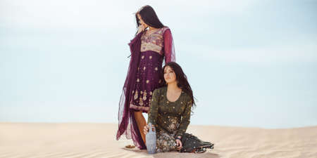 Women traveling in desert. Dehydration, overheating, thirst and heat stroke concept image with two sisters outdoors in the nature. Arabian girls lost in dunes during journey.の写真素材