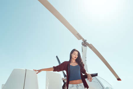 Pilot woman standing at helicopter outdoors in sunny day. Attractive young multi-racial Asian Caucasian sexy girl in jeans and shirt in airport.の写真素材