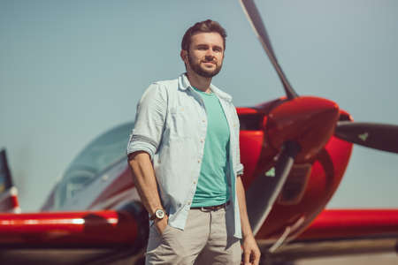 Pilot and airplane. Cheerful handsome young man pilot standing near small sport planeの写真素材
