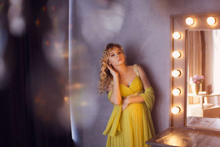 Pregnant woman. Romantic portait photo of beautiful blonde girl posing in sexy evening dress looking away at home. Indoor shot against gray wall and mirror.の写真素材