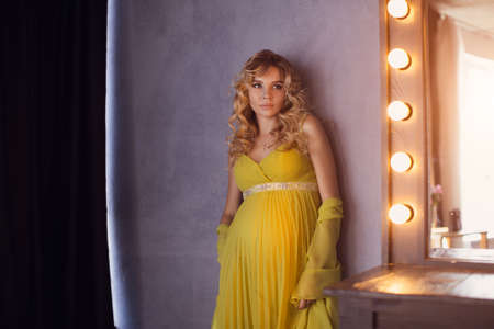 Pregnant woman. Romantic portait photo of beautiful blonde girl posing in sexy evening dress looking away at home. Indoor shot against gray wall and mirror.の写真素材