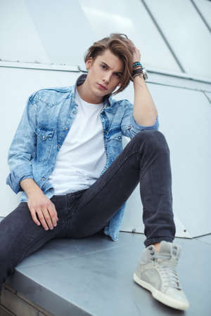 The young guy is sitting agaist business building. Poses for mens photo sessions. Beautiful appearance, clothes in rock style.の写真素材