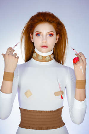 Young red hair girl holding syringes in hands pointing on her scared face with correction mark for plastic surgeryの写真素材