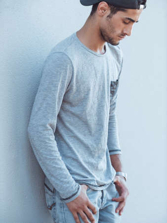 Portrait of serious young man with facial hair leaning against gray wall looking down. Handsome man wearing t-shirt baseball cap and jeans outdoorsの写真素材