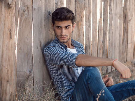 Portrait of confident young man with facial hair sitting against wooden wall looking away. Handsome man wearing shirt t-shirt and jeans resting outdoorsの写真素材