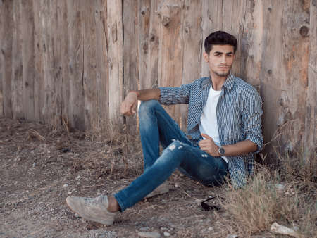 Portrait of confident young man with facial hair sitting against wooden wall looking at camera. Handsome man wearing shirt t-shirt and jeans resting outdoorsの写真素材