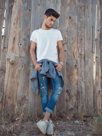 Confident young man in full length standing against wooden wall looking away. Handsome man with facial hair wearing shirt t-shirt and jeans resting outdoors.の写真素材