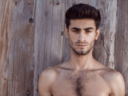 Topless young man with facial hair standing against wooden wall looking at camera. Shirtless handsome man resting outdoors.の写真素材