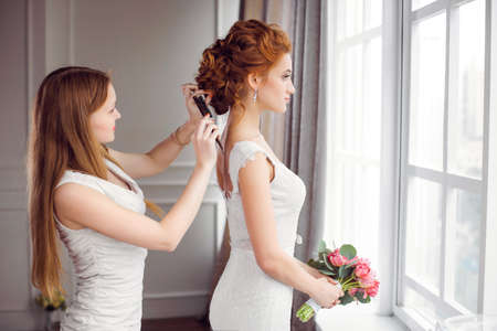 Master stylist makes the bride wedding hairstyle using hairbrush indoors at home near window....