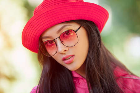 Asian woman fashion close-up portrait. Beautiful mixed race asian caucasian young girl in pink hat jaket and red sunglasses looking at camera outdoor against green blurred bokeh nature background. Gorgeous slim model in trendy casual colourful clothes posing outside in city park.の写真素材