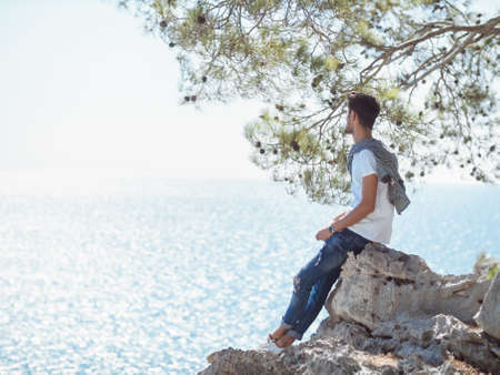 Traveler on the rocks near the sea looking far away at horizon. Summer Travel Vacation. Handsome young caucasian tourist man with sunglasses in hand in casual clothes outdoors on the nature.の写真素材