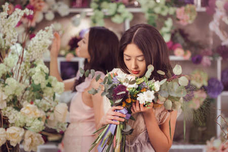 Beautiful asian florist girls making bouquet of flowers for sale against floral bokeh background in flower shop indoors. Two attractive asian females florists working in retail store. 2 playful fashion models in tender dresses posing playing with flowers.の写真素材