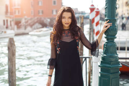 Attractive young romantic woman standing on the pier against beautiful view on venetian chanal with boats and gondolas in Venice, Italy. Travel tourist girl on vacation walking happy by Grand Canal. Gorgeous mixed race Asian Caucasian female in fashion black sexy dress smiling enjoying traveling outdoors during holidays in Europe. Model looking at camera.の写真素材