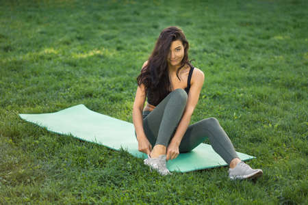 Pretty young woman sitting on yoga mat outdoors and tying shoelaces on sneakers and looking at camera. Healthy lifestyle concept.の写真素材