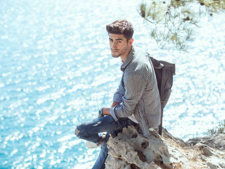 Traveler with backpack on the rocks near the sea looking at camera. Summer Travel Vacation. Handsome young caucasian tourist man in casual clothes outdoors on the nature.の写真素材
