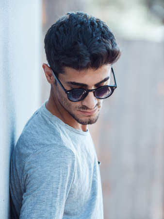 Portrait of serious young man with facial hair leaning against gray wall looking down. Handsome man...