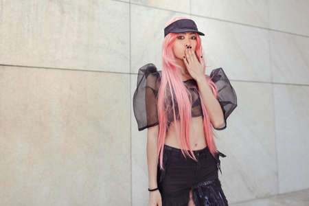 Surprised woman. Colorful portrait of young attractive asian woman in sexy dress with creative make-up and wearing pink wig outdoors. Japan anime cosplay. Trendy Japanese girl standing outside on city street against moder building.の写真素材