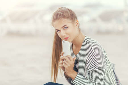 Woman with a smartphone on the beach outside. Vacation and technology. Cute stylish happy girl in casual clothes sitting on a beach near sea using the phone outdoors. Attractive caucasian model looking at cameraの写真素材