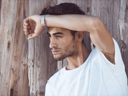 Portrait of confident young man with facial hair standing against wooden wall looking away. Handsome man wearing t-shirt outdoorsの写真素材