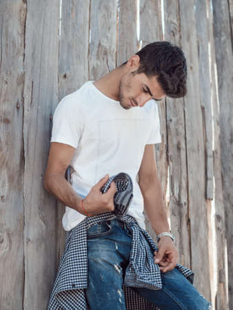 Confident young man in full length standing against wooden wall looking away. Handsome man with facial hair wearing shirt t-shirt and jeans resting outdoors.の写真素材