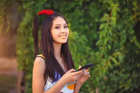 Happy young Asian woman with smartphone standing outdoors in city park. . Young Asian casual girl using her mobile phone app 4g data while relaxing on summer vacations. Fashion photo of gorgeous stylish model outsideの写真素材