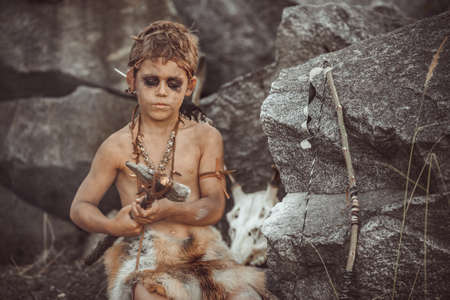 Caveman, manly boy making primitive stone weapon. Funny young primitive boy outdoors near bonfire. Evolution survival concept. Calm boy outside sitting at his rocky settlement. Prehistoric tribal man outside on wild nature. Primitive ice age man in animal skin in forest. Creative art fantasy photoの写真素材