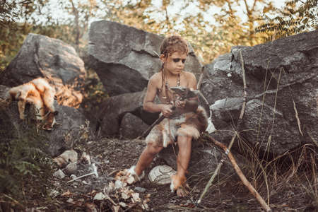 Caveman, manly boy making primitive stone weapon. Funny young primitive boy outdoors near bonfire. Evolution survival concept. Calm boy outside sitting at his rocky settlement. Prehistoric tribal man outside on wild nature. Primitive ice age man in animal skin in forest. Creative art fantasy photoの写真素材
