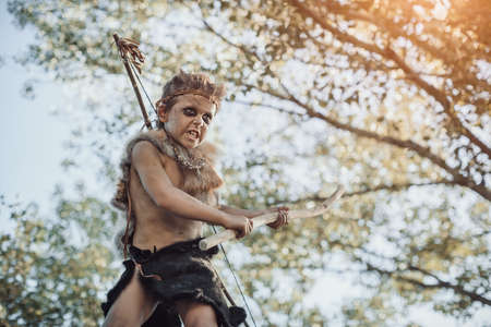 Caveman, manly boy with weapon aggressively shouting. Dramatic action photo of young primitive boy outdoors in forest.の写真素材