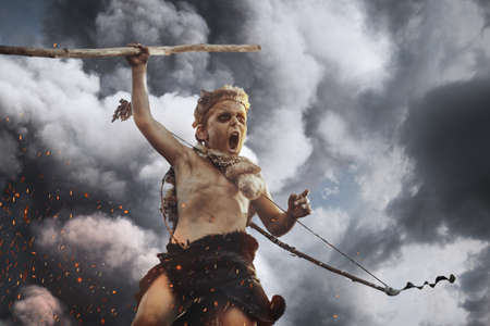 Caveman, manly boy with weapon aggressively shouting. Dramatic action photo of young primitive boy outdoors agaisnt dark clouds. Evolution survival concept. Calm boy outside standing in attack pose. Prehistoric tribal man outside on wild nature. Primitive ice age man in animal skin with bow and staff. Creative art fantasy photo.の写真素材
