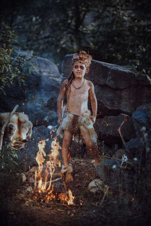 Caveman, manly boy at the fire. Scary young primitive boy outdoors near bonfire.