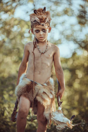 Angry caveman, manly boy with stone axe and animal skull. Prehistoric tribal boy outdoors on nature. Young shaggy and dirty savage, warrior and hunter with weapon. Primitive ice age man in animal skin. Reconstruction of Neanderthal and cro-magnon lifeの写真素材