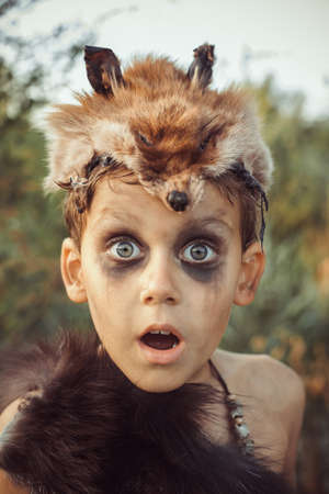 Surprised funny boy portrait. Little caveman with open mouth. Prehistoric tribal boy outdoors on nature. Young shaggy and dirty savage, warrior and hunter face. Primitive ice age man in animal skin close-up.の写真素材