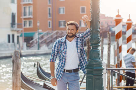 Handsome tourist man standing on the pier against beautiful view on venetian chanal with boats and gondolas in Venice, Italy. Young Caucasian male in casual clothes smiling enjoying traveling outdoors during holidays in Europe.の写真素材