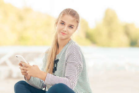 Woman with a smartphone on the beach outside. Vacation and technology. Cute stylish happy girl in casual clothes sitting on a beach near sea using the phone outdoors. Attractive caucasian model looking at cameraの写真素材