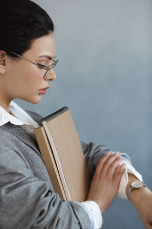 Beautiful business woman holding folder with mock up area and looking at watch standing in the office. Business concept - side view portrait photo of serious smart businesswoman in glasses casual stylish suite indoors against copy space modern gray background. Successful confidenceの写真素材