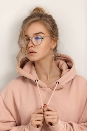 Hipster young girl over wall. Portrait of a cute positive seductive fashion model in street style posing indoors. Beautiful sensual blond woman wearing pink hoody and stylish glasses standing and looking away agaist light gray background.の写真素材