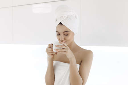 Young woman after bath or shower enjoying coffee indoors at home. Young Mixed race Caucasian Asian woman with perfect makeup in white towel with cup of coffe or tea on modern light kitchen.の写真素材