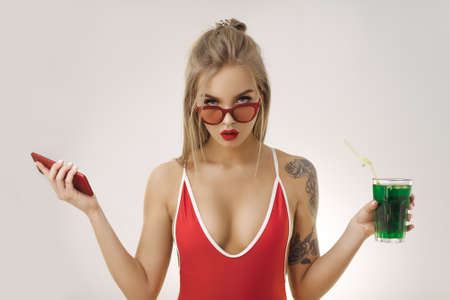 Confused woman with perfect makeup wearing sexy red bikini and retro sunglasses holding cocktail and smartphone on bright copy space background in studio. Picture of gorgeous woman standing and looking at the camera in stylish swimsuit.の写真素材