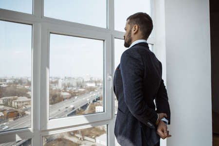 Businessman standing next to the window, looking out. Portrait of a confident brutal man in a trendy suit thinking about business indoors in office. Side view.の写真素材