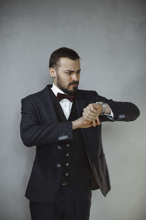 We have no more time. Portrait of the handsome serious bearded young man in trendy suit black vest and white shirt, standing, looking at the watch with worried facial expression. Indoor shot, isolated on grey background. Business and time management concept.の写真素材