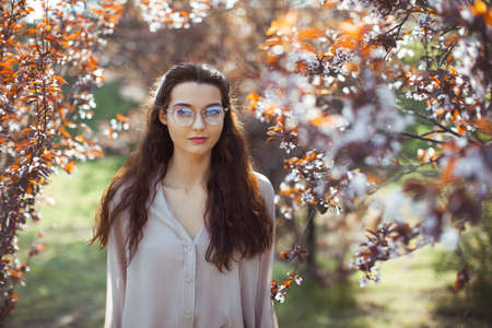 Stylish romantic creative art portrait outdoors. Beautiful mixed race Asian Caucasian young girl perfect skin tender make-up wearing glasses skirt and blouse standing against flower background.の写真素材