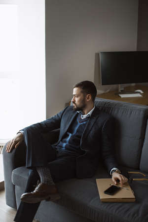 Confident young man in a trendy suit looking away while sitting indoors on the sofa. Close-up portrait of a bearded attractive gentleman in an elegant stylish clothes.の写真素材