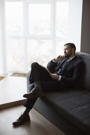 Confident concentrated handsome bearded businessman in a trendy suit writes down important ideas in a notebook sitting on a sofa in the office. Absorbedly thinking. Serious man in business wears working.の写真素材