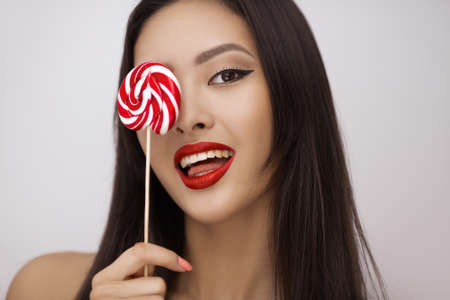 Portrait of seductive Asian woman licking colorful sugar candy with pleasure, isolated on bright studio copy space background.の写真素材