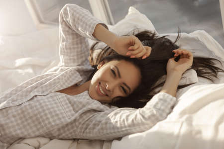 Happy woman stretching in bed in the morning. Attractive Asian girl in nightgown waking up in the...