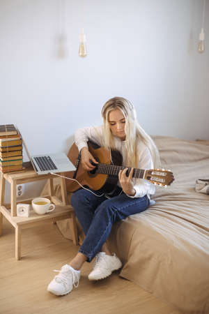 Beautiful young woman playing guitar while sitting on bed at home. Portrait of cute girl in casual clothes wearing headphones at guitar practice at home.の写真素材