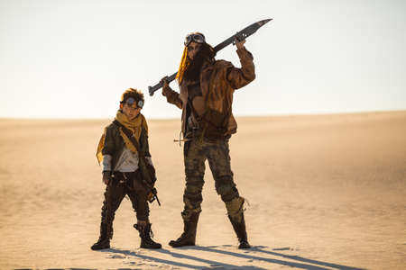 Post-apocalyptic woman and boy with weapons outdoors. Desert and dead wasteland on the background.の写真素材